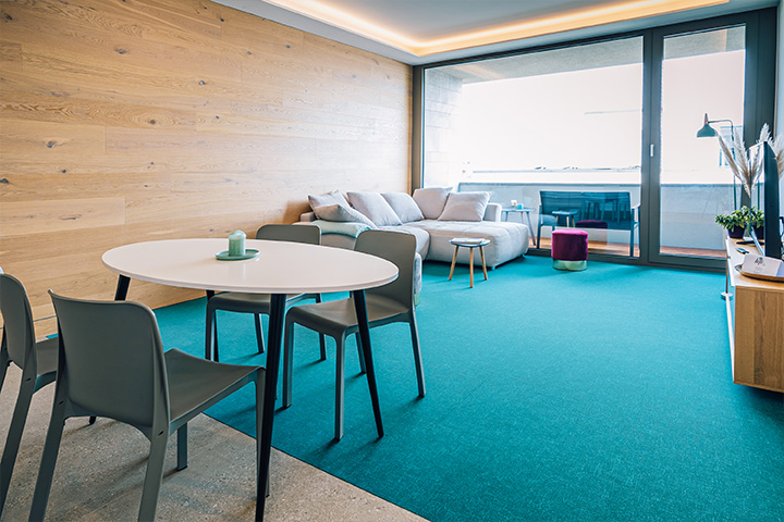 ALTUS SLEEP Apartments Haus 14: Blick in eines der Wohnzimmer mit grauer Couch vor einer holzvertäfelten Wand, petrolfarbenem Teppich, im Vordergrund ein Esstisch mit 4 Stühlen.