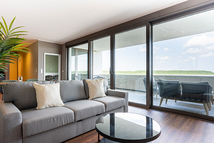 ALTUS SLEEP Apartments Haus 6: Blick in eines der Wohnzimmer mit grauer Couch, Holzfußboden, schwarzem Couchtisch und großer Terrasse im Hintergrund.