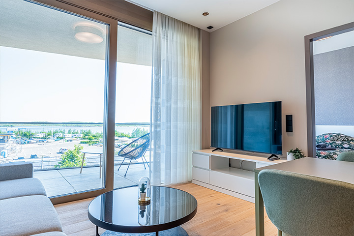 ALTUS SLEEP Apartments Haus 8: Blick in eines der Wohnzimmer mit Holzfußboden, TV-Bank mit Flat-TV, schwarzem Couchtisch und Blick auf den See im Hintergrund.