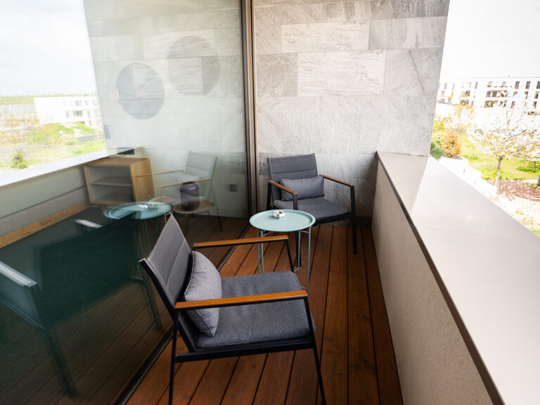 Das Bild zeigt den Balkon in Apartment 1 in Haus 14 (LAKESIDE). Braune Dielen, zwei bequeme Sessel und ein Beistelltisch mit Aschenbecher.