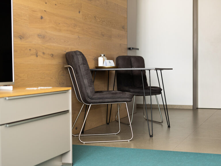 Das Bild zeigt die Essecke in Apartment 1 in Haus 14 (LAKESIDE). helle Fliesen, holzvertäfelte Wand, dunkler Tisch und zwei Stühle mit dunkelgrauem Polster.