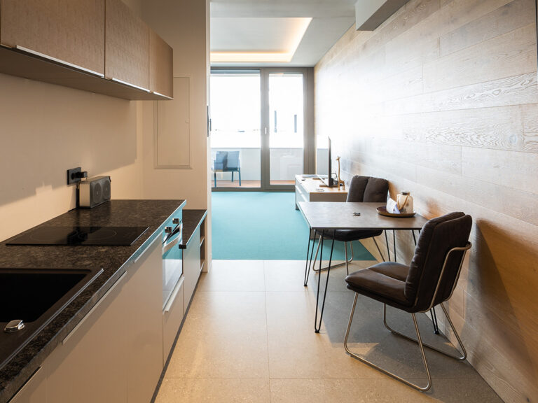 Das Bild zeigt das Apartment 1 in Haus 14 (LAKESIDE). Vorn Küche mit hellen Fliesen, grauer Küchenfront, 2 Stühlen und Tisch, im Hintergrund petrolfarbener Teppich, große bodentiefe Fenster mit Balkontür.