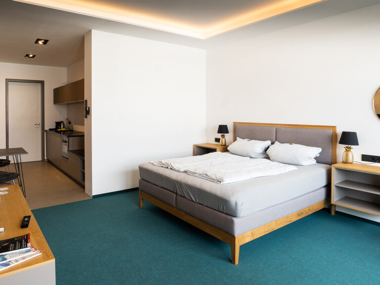 Das Bild zeigt das Schlafzimmer in Apartment 1 in Haus 14 (LAKESIDE). Petrolfarbener Teppich, helle Wände, Doppelbett mit gepolstertem Kopfteil.