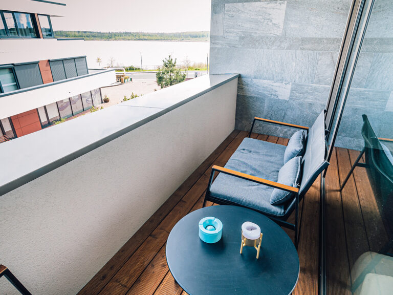 Das Bild zeigt den Balkon in Apartment 2 in Haus 14. Outdoor Sofa sowie ein kleiner Beistelltisch mit Aschenbecher, im Hintergrund ein Teil des Nachbargebäudes sowie der Zwenkauer See.