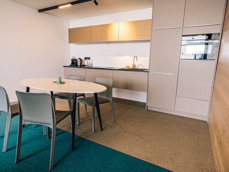 Das Bild zeigt die offene Küche in Apartment 2. Graue und goldene Fronten, helle Bodenfließen, Tisch und Stühle.