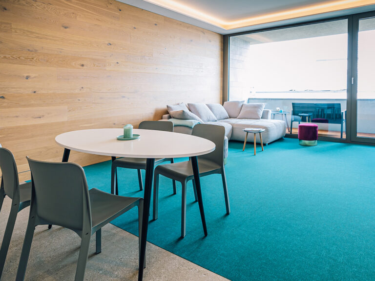 Das Bild zeigt den Wohn- und Essbereich in Apartment 2. Holzvertäfelte Wand, petrolfarbener Teppich, im Vordergrund Tisch und Stühle, im Hintergrund Sofa und Balkon.