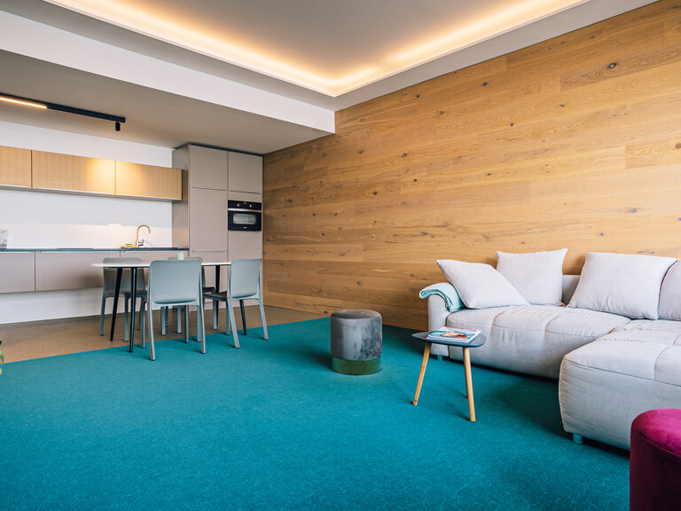 Das Bild zeigt den Wohn- und Essbereich in Apartment 2. Holzvertäfelte Wand, petrolfarbener Teppich, im Hintergrund Küchenzeile, Tisch und Stühle, im Vordergrund Sofa vor holzvertäfelter Wand.