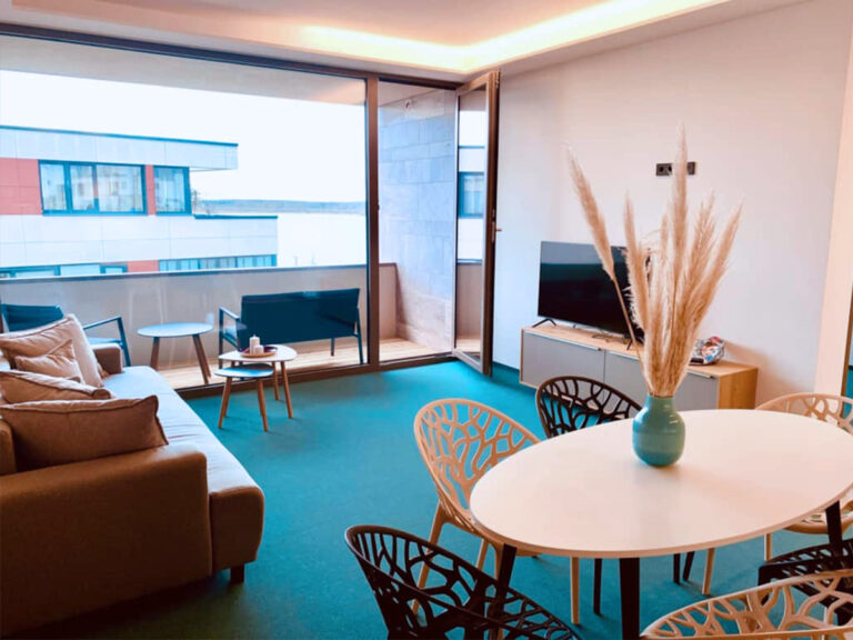 Das Bild zeigt den Wohn- und Essbereich in Apartment 3. Im Vordergrund Esstisch und Stühle, dahinter Sideboard mit Flachbild-TV, Sofa und möblierter Balkon.