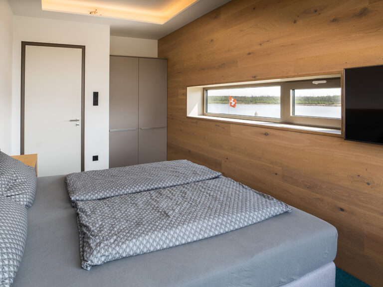 Das Bild zeigt dasSchlafzimmer in Apartment 4. Holzvertäfelte Wand mit Fenster zum See sowie Flachbild-TV, Kleiderschränke mit grauer Front, Doppeltbett.