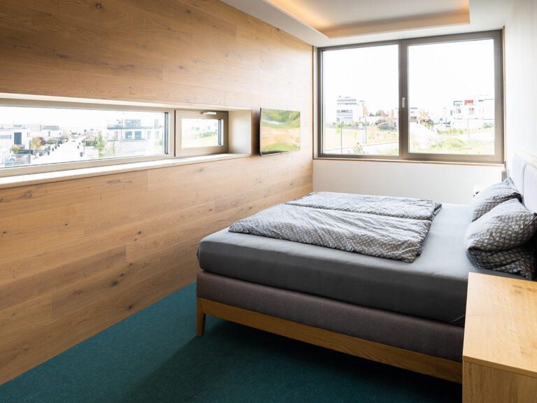 Das Bild zeigt das Schlafzimmer in Apartment 4. Holzvertäfelte Wand mit Fenster zum See sowie Flachbild-TV, großes Fenster zur Stadt, Doppeltbett.