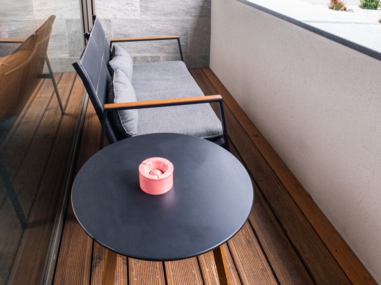 Das Bild zeigt den Balkon in Apartment 5. Braune Dielen, gepolstertes Sofa, Beistelltisch mit Aschenbecher.
