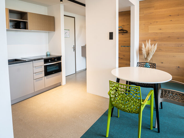 Das Bild zeigt die offene Küche in Apartment 5. Küchenzeile mit E-Herd, Sitzgruppe und im Hintergrund Eingang mit Garderobe.