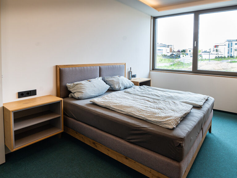 Das Bild zeigt das Schlafzimmer in Apartment 5. Doppelbett mit gepolstertem Kopfteil, Nachtschränkchen, im Hintergrund großes Fenster mit Landblick.