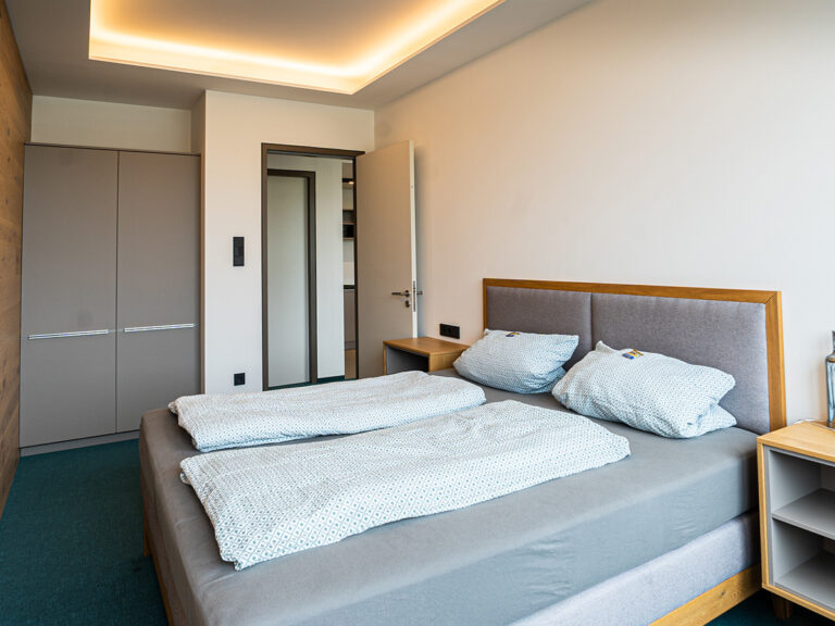 Das Bild zeigt das Schlafzimmer in Apartment 5. In der Ecke hoher Kleiderschrank, davor Doppelbett mit gepolstertem Kopfteil, Im Hintergrund Tür zum Flur.