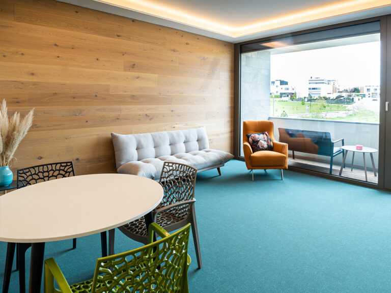 Das Bild zeigt den Wohn- und Essbereich in Apartment 5. Holzvertäfelte Wand, petrolfarbener Teppich, hellgraues Sofa und orangefarbener Sessel, Tischgruppe. Im Hintergrund Balkon mit Sitzbank.