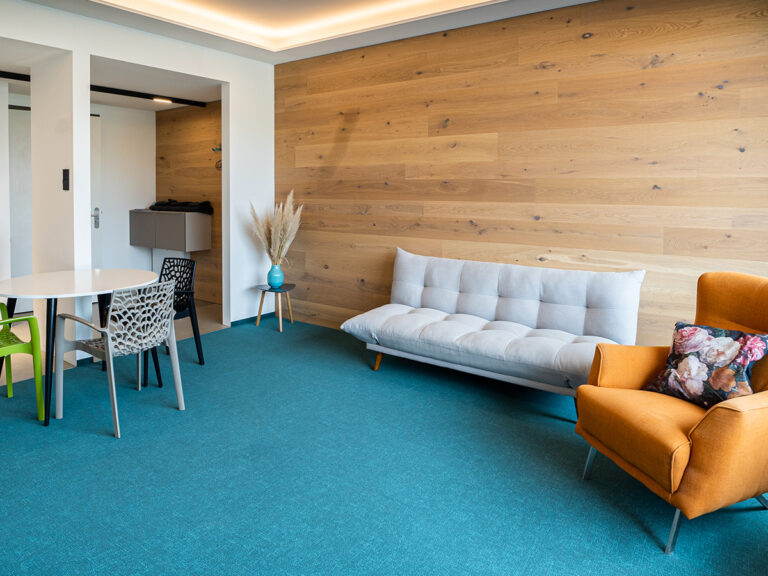 Das Bild zeigt den Wohn- und Essbereich in Apartment 5. Holzvertäfelte Wand, petrolfarbener Teppich, hellgraues Sofa und orangefarbener Sessel, Tischgruppe. Im Hintergrund Eingang mit Garderobe.