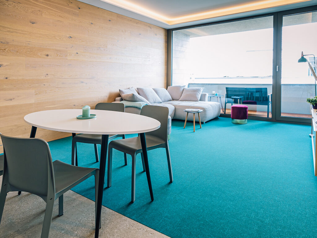 Haus 14: Blick in eines der Wohnzimmer mit grauer Couch vor einer holzvertäfelten Wand, petrolfarbenem Teppich, im Vordergrund ein Esstisch mit 4 Stühlen.