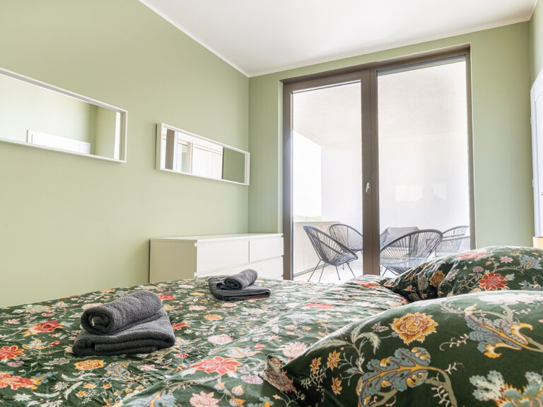 Nahaufnahme eines Bettes mit hellgrauem Bettlaken und grün-gemusterter Bettwäsche, im Hintergrund eine grüne Wand, Spiegel, Kommode und Loggia.