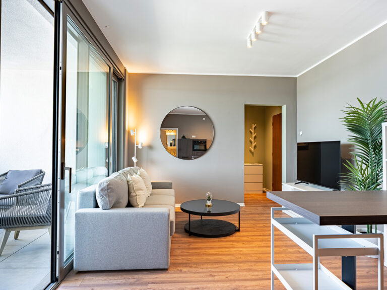 Das Bild zeigt das Wohnzimmer in einem Haus-6-Apartment, mit grauer Couch und Loggia im Hintergrund.