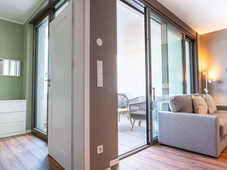 Das Bild zeigt das Wohnzimmer in Apartment 2 bzw. 6 in Haus 6, mit grauer Couch und Loggia im Hintergrund.