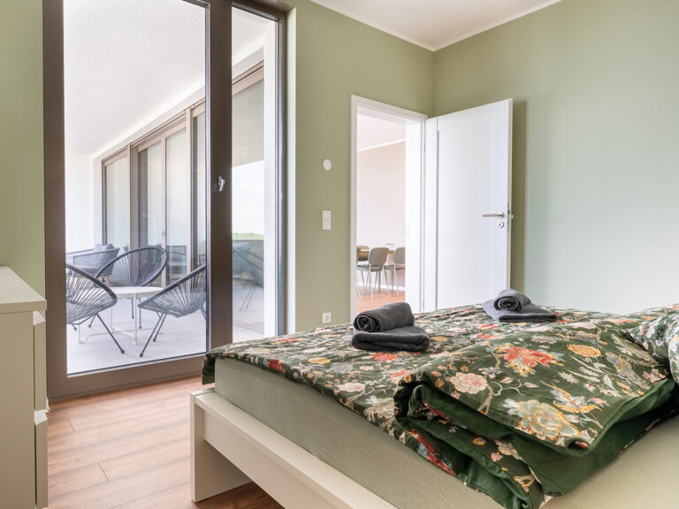 Schlafzimmer in Apartment 3 und 7 mit weißem Bett, grüner Bettwäsche, im Hintergrund großes Fenster zur Loggia.