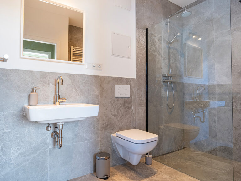 Das Bild zeigt eines der Bäder in Haus 6 mit grauen Fliesen, ebenerdiger Dusche, WC und Toiletteneimer.