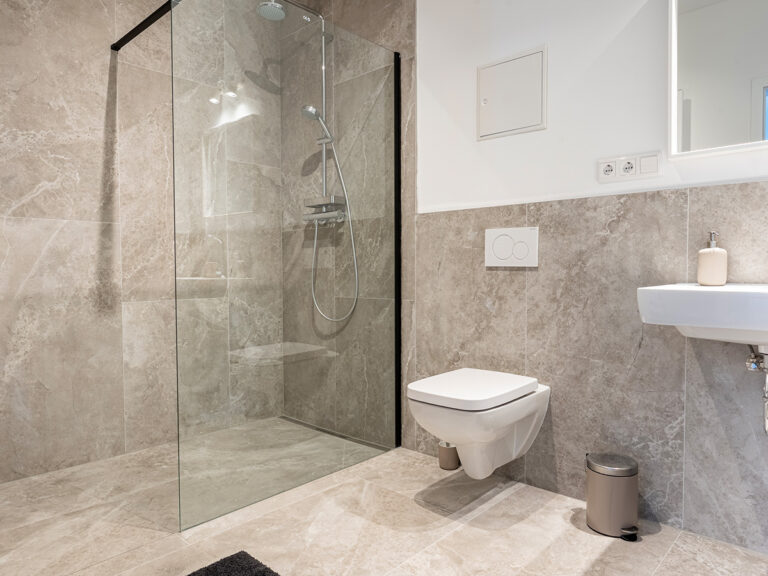Das Bild zeigt eines der Bäder in Haus 6 mit grauen Fliesen, ebenerdiger Dusche, WC und Toiletteneimer.