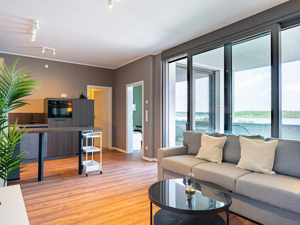Das Bild zeigt das Wohnzimmer in einem Haus-6-Apartment, mit grauer Couch, Küchenecke und Loggia im Hintergrund.