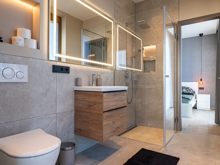 Bad in Apartment 2 in Haus 8: graue Fliesen, WC, beleuchteter Spiegel, Waschbecken und ebenerdige Dusche im Hintergrund.
