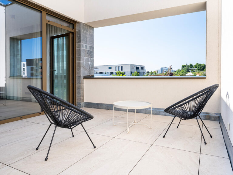 Dachterrasse in Apartment 2 in Haus 8: helle Fliesen, weißer Couchtisch, 2 schwarze Outdoor-Sessel; Blick ins Landesinnere.
