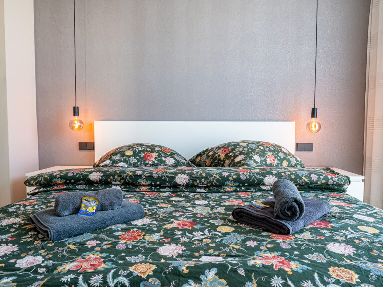 Schlafzimmer in Haus 8: Holzfußboden, graue Tapete, weißes Doppelbett.