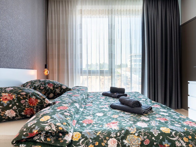 Schlafzimmer in Apartment 2 in Haus 8: Holzfußboden, graue Tapete, weißes Doppelbett, große Fenster mit Vorhängen.