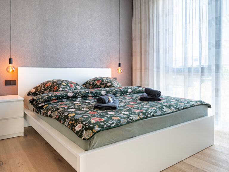 Schlafzimmer in Apartment 2 in Haus 8: Holzfußboden, graue Tapete, weißes Doppelbett, große Fenster mit hellen Vorhängen.