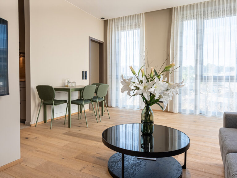 Wohnbereich in Apartment 2 in Haus 8: Holzfußboden, schwarzer Couchtisch, grüne Sitzgruppe, große Fenster mit hellen Vorhängen.