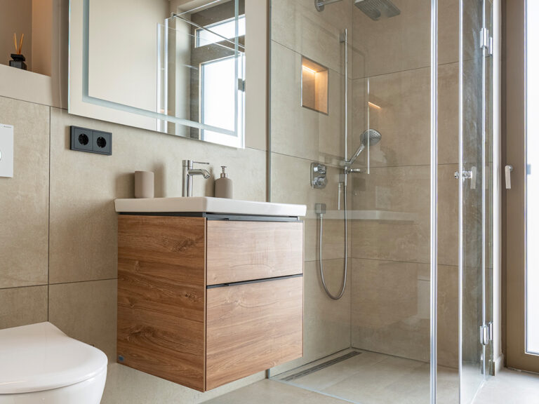 Bad in Apartment 3 in Haus 8: helle Fliesen, WC und Waschbecken, beleuchteter Spiegel, im Hintergrund ebenerdige Dusche.