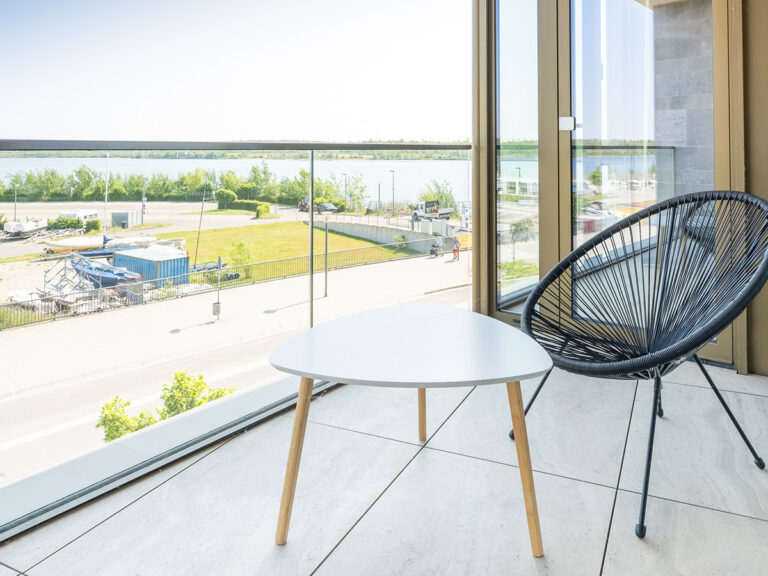 Balkon in Apartment 4 in Haus 8: helle Fliesen, weißer Tisch, schwarzer Outdoor-Sessel, im Hintergrund See.