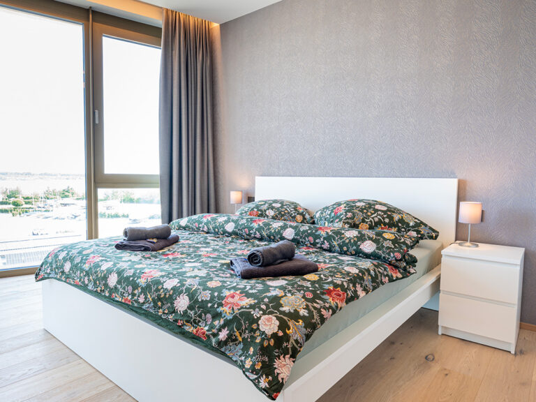 Schlafzimmer in Apartment 4 in Haus 8: Holzfußboden, graue Tapete, weißes Bett, im Hintergrund große bodentiefe Fenster mit Seeblick.